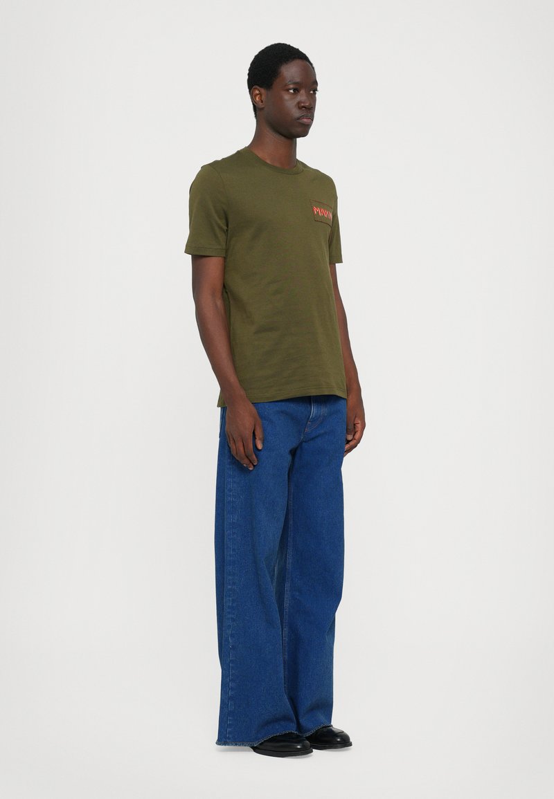 Camiseta de algodón verde oliva con mangas cortas y un bolsillo frontal con bordado rojo, combinada con unos vaqueros de denim azul de pierna ancha.