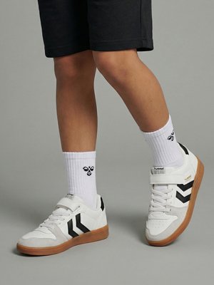 Kind draagt witte sneakers met zwarte chevronstrepen en gumzolen, gecombineerd met witte crew-sokken en zwarte korte broeken op een grijze achtergrond.