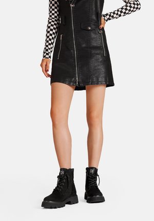 Mini abito in pelle nera con dettagli a zip e tasche, abbinato a stivali militari neri con lacci, caratterizzati da accenti testurizzati e suole spesse.