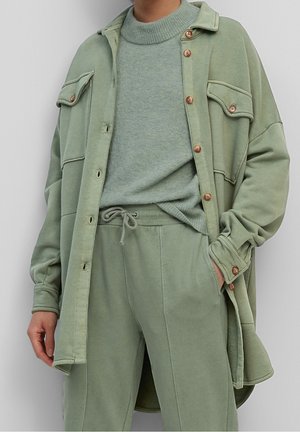 Person wearing sage green layered outfit: knit sweater, oversized button-up jacket with flap pockets, and drawstring pants, hand partially in pocket.