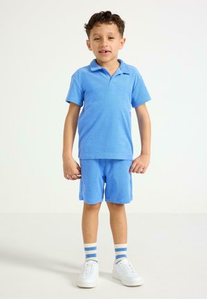 Ung dreng står, iført en blå poloshirt med korte ærmer, matchende shorts, stribede sokker og hvide sneakers mod en ensfarvet baggrund.