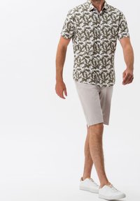Muž jde a má na sobě košili s krátkým rukávem s vzorem listů, béžové šortky a bílé tenisky na bílém pozadí.