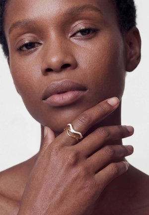 Nahaufnahme einer Person mit kurzen Haaren, die ihr Gesicht berührt und am Zeigefinger einen modernen Ring in Gold- und Weißdesign trägt.