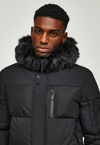 Schwarze Daunenjacke mit einem mit Fell gefütterten Kragen, ausgestattet mit Reißverschlusstaschen und einer gesteppte Textur. Vorderer Verschluss mit Druckknöpfen.