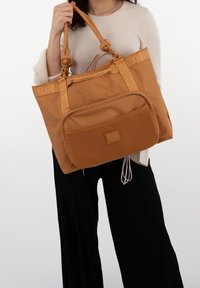 Person som håller en stor beige tygväska med ett meshnätfickor och två knutna handtag, iförd en beige topp och svarta byxor.