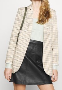 Beige kostkovaný blazer s zelenými akcenty a dlouhými rukávy, kombinovaný s černou koženou sukní ve tvaru A. V designu je viditelná texturovaná látka.