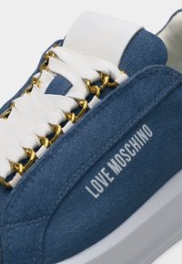 Μπλε τζιν αθλητικό παπούτσι Love Moschino με λευκά κορδόνια που περνούν μέσα από χρυσές θηλιές αλυσίδας και λευκή καουτσούκ σόλα.