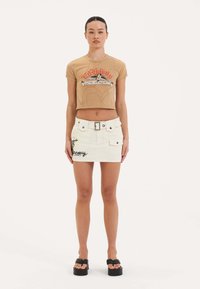 Beige cropped t-shirt med grafisk tryk, parret med en hvid denim miniskirt med lommer, bælte og broderede detaljer.