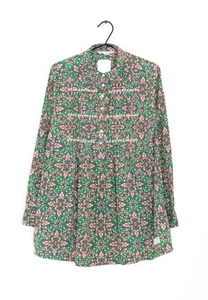 Camisa de manga larga con botones, con un patrón geométrico floral en verde y rosa, colgada en una percha negra.