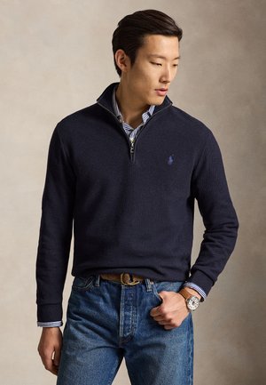 Mand iført navy kvart-zip sweater over stribet skjorte, blå jeans og brun læderur, kigger ned med den ene hånd i lommen.