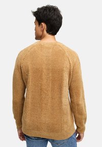 Mand iført en struktureret tan farvet kabelstrik sweater og blå jeans, set fra ryggen mod en helt hvid baggrund.