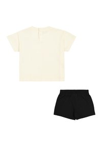 Camicia leggera a maniche corte di colore crema con bottoni sul retro, abbinata a pantaloni corti con cintura elastica nera, entrambi realizzati in tessuto morbido.