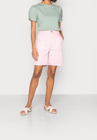 Donna che indossa una blusa a maniche puff verde chiaro, pantaloni corti rosa a vita alta e sandali bianchi incrociati, in piedi davanti a uno sfondo semplice.