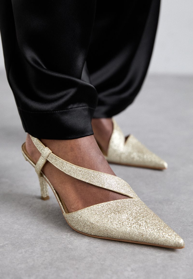 Kuldsed glitterslingback kontsad, millel on terav nina ja peen konts, risti minevad rihmad ja sile, tekstuuriga pind.