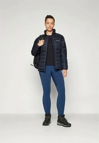 Mladostnik nosi črno puhovko, modre legice in črne pohodne čevlje, na eni rami nosi črn nahrbtnik.