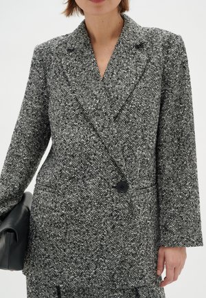 Vrouw draagt een zwart-witte blazer met visgraatmotief, brede revers, enkele knoop en houdt een zwarte clutch vast.