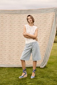 Femme debout, les bras croisés, vêtue d’un haut en tricot blanc sans manches, d’un short en denim longueur genou et de sandales compensées bleues espadrilles sur l’herbe devant un tissu à motif.