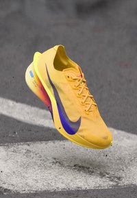Yellow Nike running shoe with purple swoosh, mid-air above asphalt and white road markings, small debris beneath the sole.