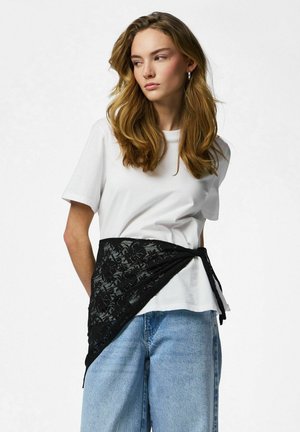 Jonge vrouw met lang golvend haar, gekleed in een wit T-shirt, lichtblauwe spijkerbroek en een zwart kanten sjaaltje om haar middel gestrikt.