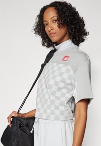 Tricou scurt gri cu un model de tablă de șah, guler tip funnel și o etichetă roșie. Modelul ține o geantă neagră și poartă pantaloni deschisi la culoare.