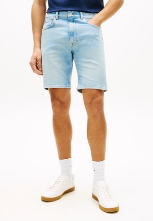 Mann trägt hellblaue Jeansshorts, weiße Sportsocken und weiße Sneaker mit braunen Sohlen, Hände in den Taschen, vor weißem Hintergrund.