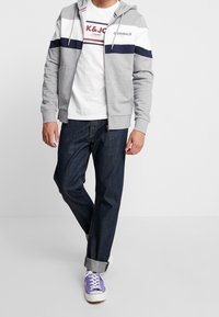 Sudadera con capucha gris con cremallera y rayas azul marino, usada sobre una camiseta blanca con texto. Jeans de mezclilla oscura y zapatillas moradas completan el look.