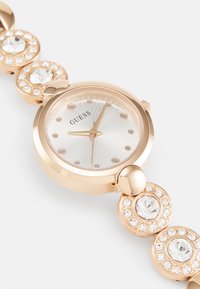 Gold-tone watch with a round face, white patterned dial, crystal hour markers, and linked band featuring circular accents with embedded rhinestones.
