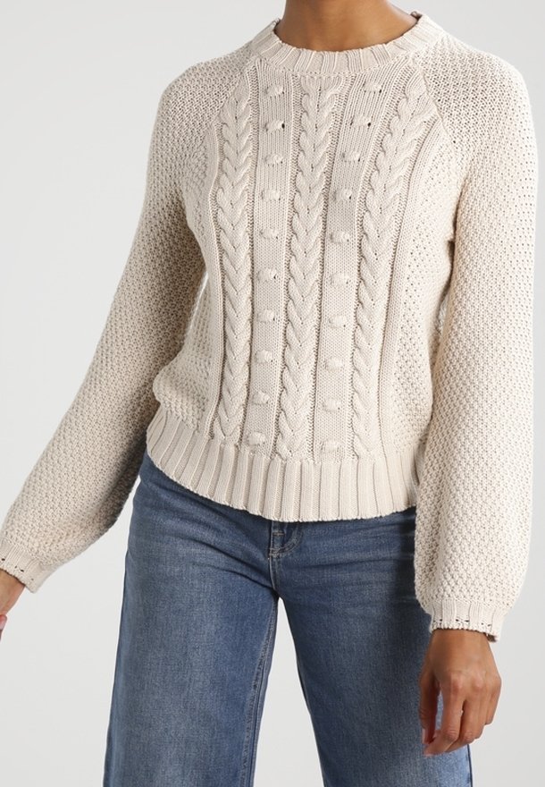 Beige Grobstrickpullover mit Muster aus Noppen und strukturierten Stichen, langen Puffärmeln, kombiniert mit mittelblauen Jeans.