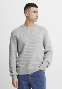 Pulover gri deschis, dintr-un material texturat cu un model în diamant, cu decolteu rotund și manșete și tiv cu nervuri, purtat cu blugi albaștri.