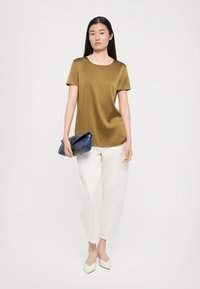 Olive green short-sleeve top, smooth texture; paired with white loose-fitting trousers and a navy clutch bag; model poses upright.