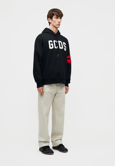 Ung mann står, iført en svart GCDS-hettegenser med røde striper på ermene, beige bukser og svarte joggesko mot en ensfarget hvit bakgrunn.