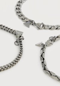 Trois bracelets en chaînes métalliques argentées avec différents styles de maillons, chacun équipé d'un fermoir à mousqueton et d'un petit pendentif triangulaire.