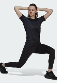 Camiseta deportiva negra con un patrón sutil, combinada con leggins y zapatillas negras. Modelo en posición de zancada, con las manos detrás de la cabeza, enfocando hacia adelante.