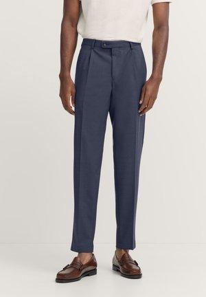 Marineblaue, maßgeschneiderte Hosen mit flachem Vorderteil, zwei Falten, Knopfverschluss und taillierter Passform. Getragen mit braunen Slippers und einem hellen Hemd.