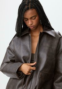 Giacca in faux leather marrone scuro con ampio colletto, chiusura frontale con zip e tasca sul petto. La superficie ha una finitura leggermente strutturata.