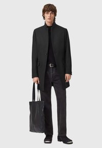 Mustapukuinen takki, jossa on korkea kaulus ja yksittäinen nappikiinnitys, yhdistettynä mustaan poolopaitaan, tummaan farkkuihin ja tekstuuriin mustaan kantokassiin.