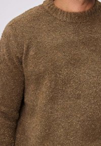 Pull en laine brune texturée porté sur le torse et le cou d'une personne, avec un col rond.