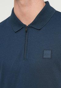 Μπλε ναυτικό πόλο με ζιπ νέκλα και μικρό τετράγωνο λογότυπο BOSS στο αριστερό στήθος, που φοριέται από έναν άνθρωπο.