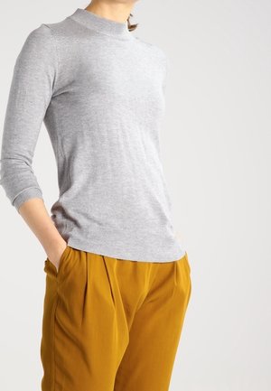 Un pull en tricot gris avec un col montant et des manches longues, associé à un pantalon plissé jaune moutarde. Le tissu semble doux et texturé.