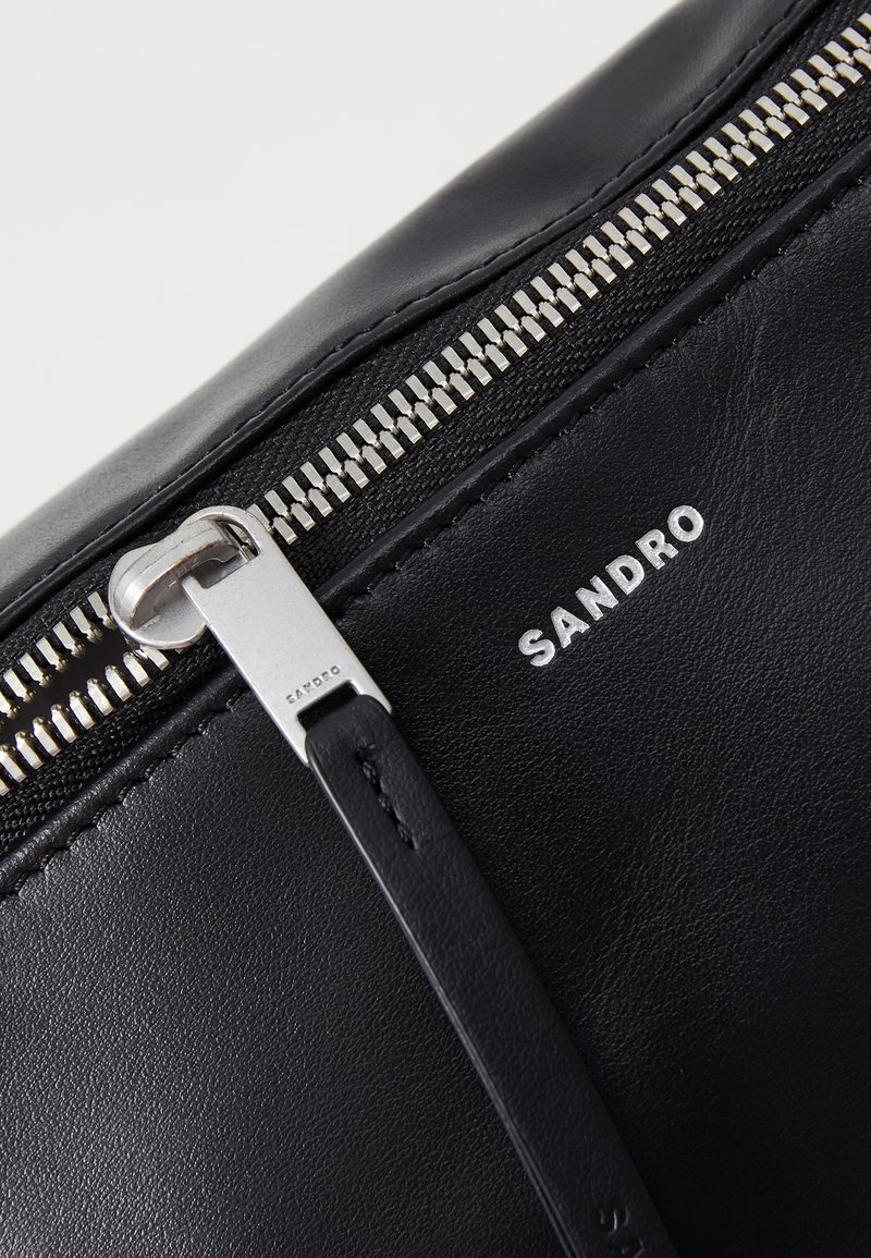 Un sac en cuir noir avec une fermeture éclaire métallique argentée, une marque imprimée "SANDRO", une texture lisse et un design minimaliste.
