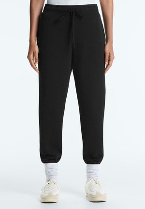 Osoba ubrana w czarne joggery z ściągaczem, białe skarpety za kostkę i białe trampki, stojąca z ramionami swobodnie opuszczonymi wzdłuż ciała.