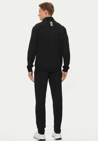 Giacca nera con zip e pantaloni abbinati realizzati in un materiale testurizzato, con un accento a righe sulla parte posteriore del colletto.