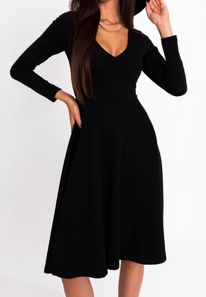 Mujer vestida con un vestido midi negro de manga larga con escote en V, complementado con cadenas doradas superpuestas, posando frente a un fondo blanco liso.