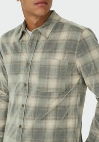Karirana flanelasta srajca v svetlo zeleni in krem barvi, z gumbom spredaj, razširjenim ovratnikom in žepom na prsi. Teksturovana tkanina.