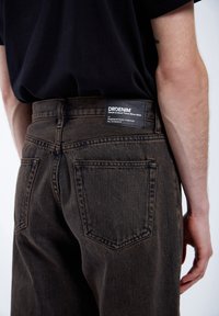 Tmavě hnědé džíny z denimu s klasickým střihem. Obsahují zadní kapsy a černou nášivku s brandingem "DRDENIM" umístěnou na pase.