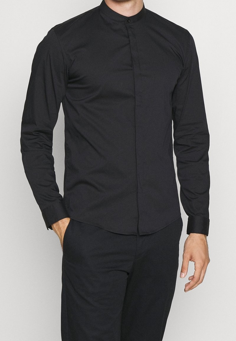 Homme portant une chemise noire ajustée à col haut et manches longues avec boutons dissimulés et pantalon noir assorti, une main dans la poche.