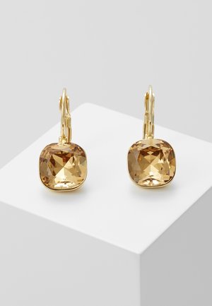 Złote kolczyki z dużymi, kwadratowo szlifowanymi bursztynowymi kamieniami, wyeksponowane na białym sześciennym podium na szarym tle.