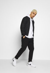 Giubbotto bomber nero sopra una felpa bianca con cappuccio, abbinato a pantaloni neri e sneakers bianche con un'accenno nero. Sfondo neutro.