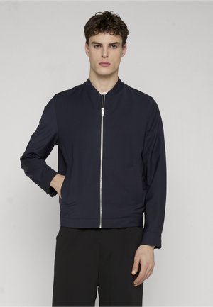 Młody mężczyzna w granatowej kurtce na zamek, z jedną ręką w kieszeni, stoi na tle gładkiego, jasnoszarego tła.