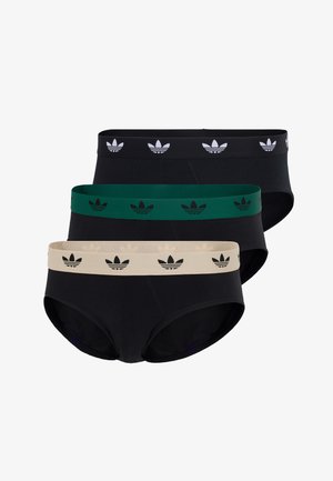 Jonge man staat op blote voeten, draagt zwarte slip met een groene tailleband met herhalend trefoil-logo, tegen een effen witte achtergrond.